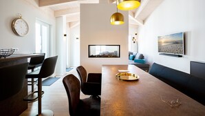 Eine moderne Küche mit Holzbalkendecke, ein Essbereich mit langem Tisch und Stühlen sowie ein Wohnzimmer mit Sofa und einem gerahmten Bild an der Wand.