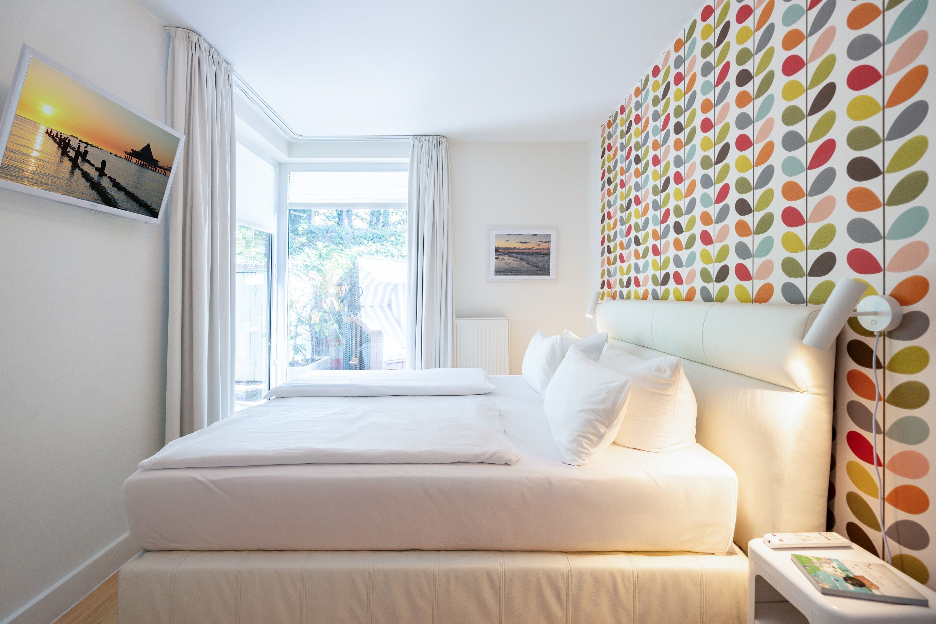 A hotel room with a large bed, a bedside table, a colorful wall art, and a window with curtains.