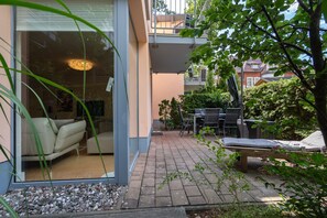 Un patio exterior moderno con puerta de cristal que da a un salón, un pequeño jardín con un banco y vistas a casas vecinas.