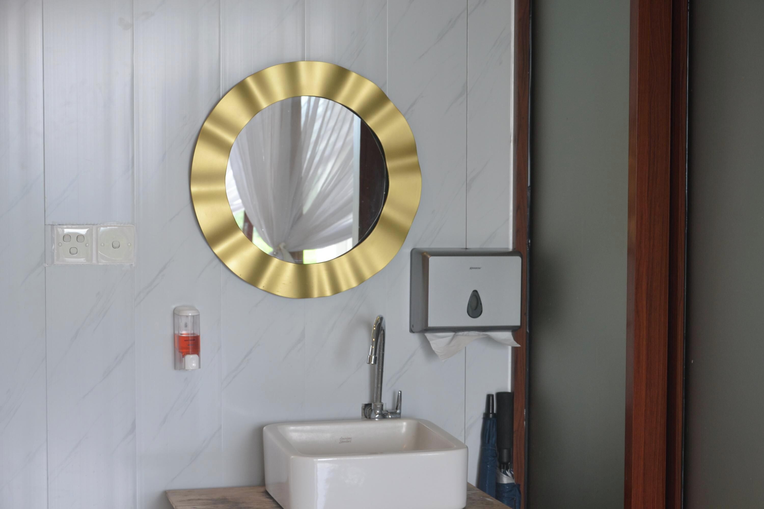 A bathroom with a white sink, a gold-framed mirror, and a soap dispenser on the wall.
