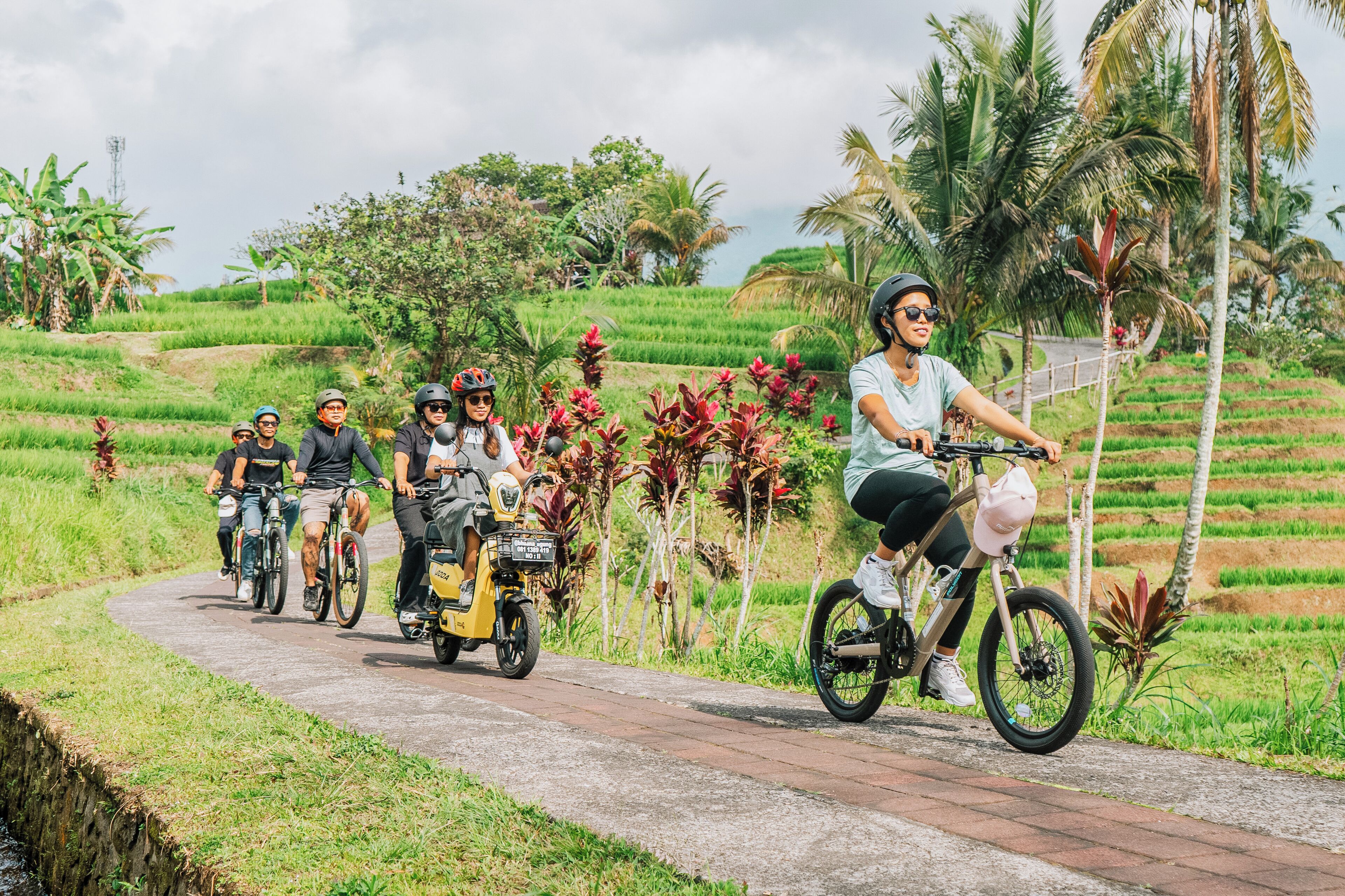 Um grupo de pessoas andando de bicicleta por um caminho pavimentado, cercado por vegetação exuberante e palmeiras.