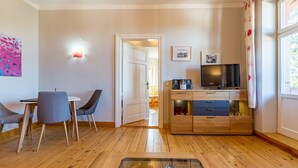 Ein modernes Wohnzimmer mit Holzboden, einem Esstisch mit Stühlen, einer TV-Konsole mit Lautsprechern und einer Tür zu einem weiteren Raum.