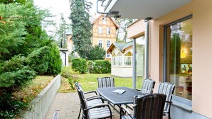 Eine Terrasse im Freien mit Tisch und Stühlen, umgeben von Pflanzen und einem Gebäude im Hintergrund.