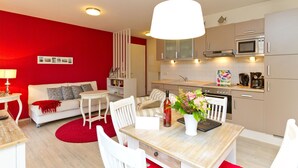 Eine moderne Küche mit Mikrowelle, Backofen und Kaffeemaschine. Ein Esstisch mit Stühlen und ein roter Teppich sind vorhanden. Ein weißes Sofa mit Kissen lehnt an einer roten Wand.