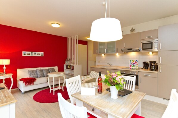 A modern kitchen with a microwave, oven, and coffee maker. There is a dining table with chairs and a red rug. A white sofa with pillows is against a red wall.