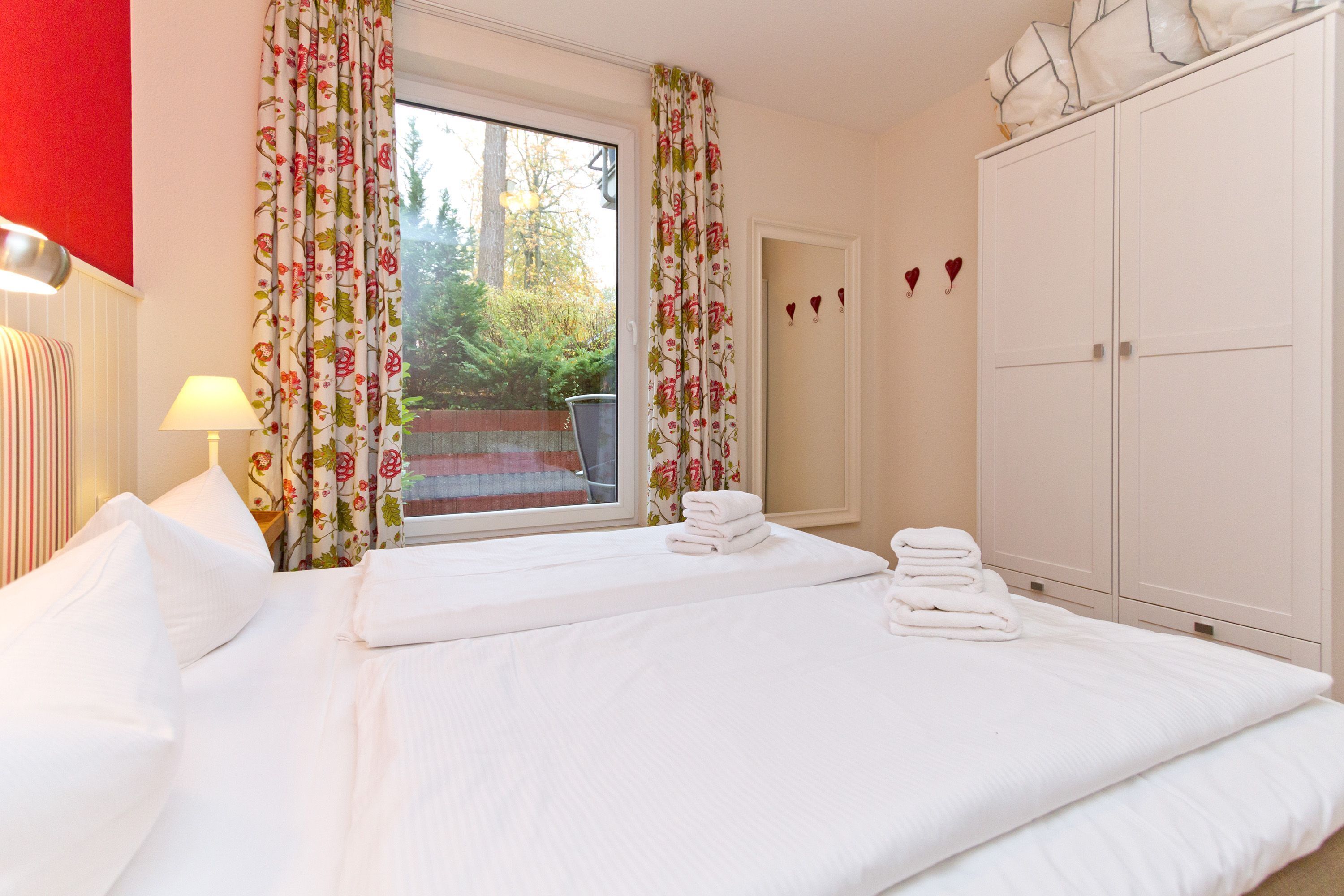 Ein Hotelzimmer mit einem großen Bett, einem Fenster mit floralem Vorhang, einem Kleiderschrank und Blick ins Grüne.