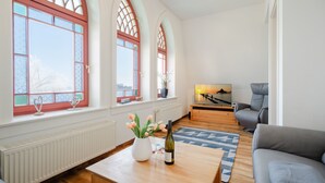 Una sala de estar con una mesa de centro de madera, un sofá y un televisor. Hay ventanas grandes con marcos rojos y vistas al exterior.
