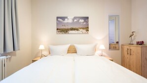 A bedroom with a large bed, two bedside lamps, a wooden headboard, a nightstand, a wooden wardrobe, a mirror, and a beach landscape painting on the wall. - Villa Medici Heringsdorf, App. 02 (Heringsdorf)
