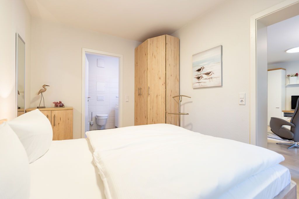 Een slaapkamer met een bed, een houten kledingkast, een bureau met stoel, en een badkamer die door een open deur zichtbaar is.