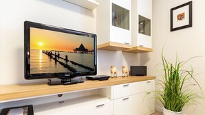 A modern living room with a flat-screen TV displaying a sunset scene, a wooden console table, white cabinetry, a small plant, and a framed picture on the wall. - Villa Medici Heringsdorf, App. 02 (Heringsdorf)