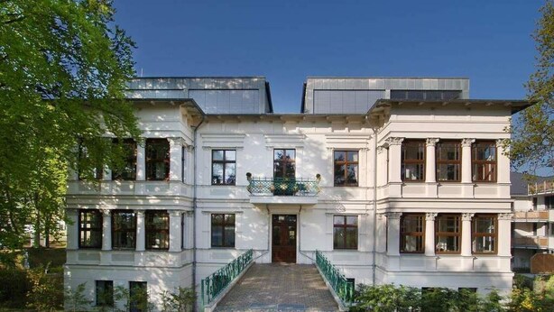 A two-story building with a central entrance, symmetrical windows, and a green railing.