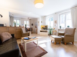 Un salon spacieux avec un canapé, une table basse et un téléviseur. Il y a également un coin repas avec une table et des chaises. La pièce est dotée de grandes fenêtres avec des rideaux.