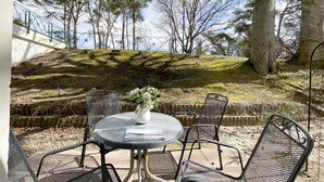 An outdoor patio with a round table and chairs, a vase with flowers, and a view of trees and a hillside.