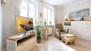 A modern living room with a flat-screen TV, a comfortable armchair, a wooden coffee table, and a view of the sea through large windows.