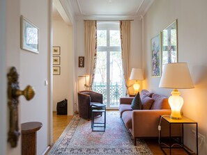 A living room with a sofa, armchair, and a table with a lamp. There is a window with curtains and a view of the outside.