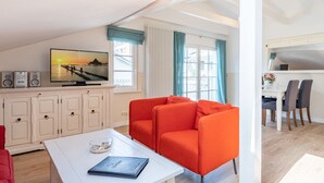 A modern living room with a white sofa, a television displaying a sunset scene, and a dining area with a white table and chairs. - Villa Hartmann-Drewitz, App. 08 (Heringsdorf)