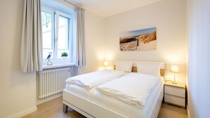 A bedroom with a bed, bedside lamps, a window with curtains, a radiator, and a painting on the wall. - Villa Medici Heringsdorf, apt. 01 (Heringsdorf)