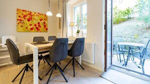 A dining area with a table and chairs, a window with a view of greenery, and a painting on the wall. - Villa Medici Heringsdorf, apt. 01 (Heringsdorf)