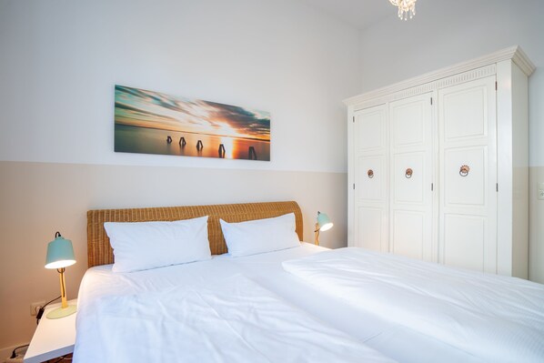 Un lit bien fait, avec du linge de lit blanc, une tête de lit en osier et deux lampes de chevet. Un grand tableau représentant un coucher de soleil est accroché au mur au-dessus du lit.