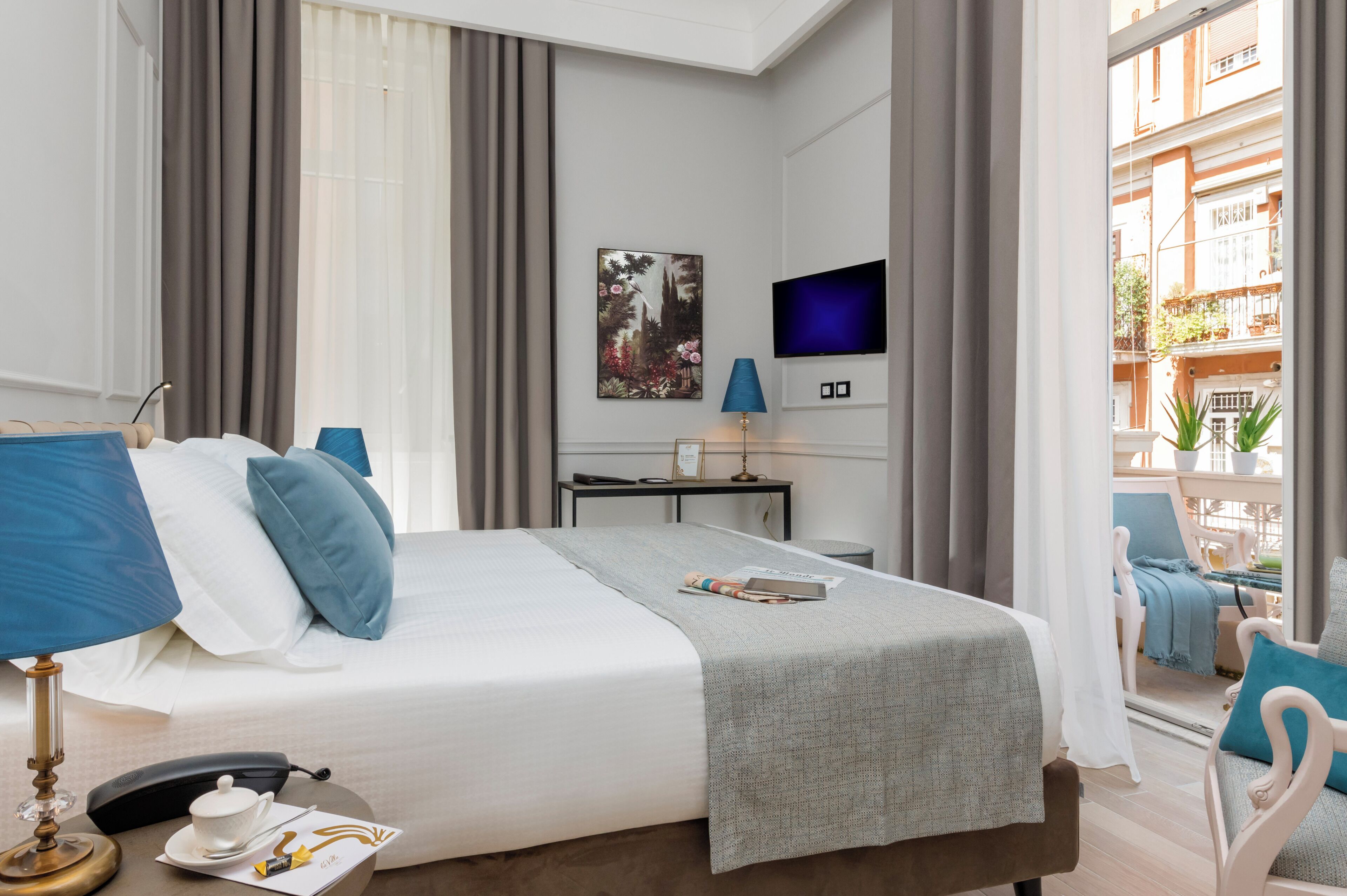 Ett hotellrum med en säng, ett skrivbord, en TV och en balkong med utemöbler.