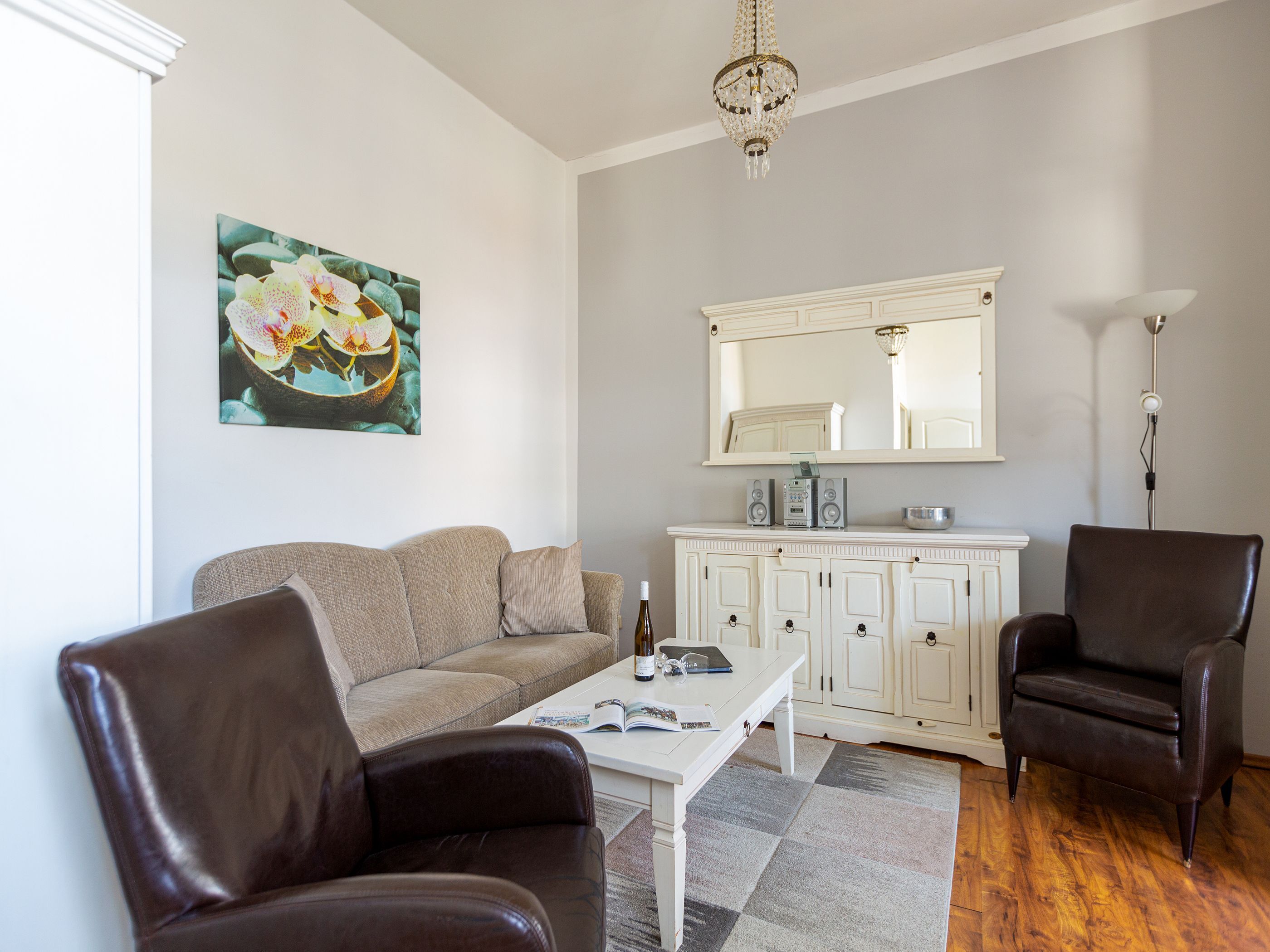 Un salon avec un canapé beige, deux fauteuils en cuir, une table basse blanche sur laquelle se trouvent un magazine et une bouteille, un meuble blanc avec des haut-parleurs, et un grand miroir au-dessus du meuble.
