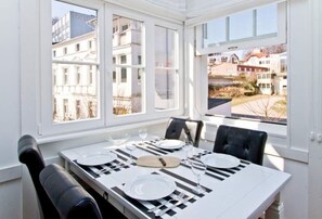 Uma área de refeições com uma mesa posta para quatro pessoas, janelas amplas e vista para edifícios.