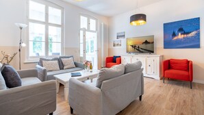 A modern living room with grey sofas, a red armchair, and a white cabinet. There are large windows and a painting on the wall. - Villa Hartmann-Drewitz, App. 06 (Heringsdorf)