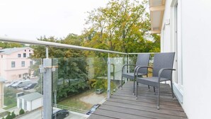 A balcony with two wicker chairs, a glass railing, and a view of a residential area with trees and a pink building.