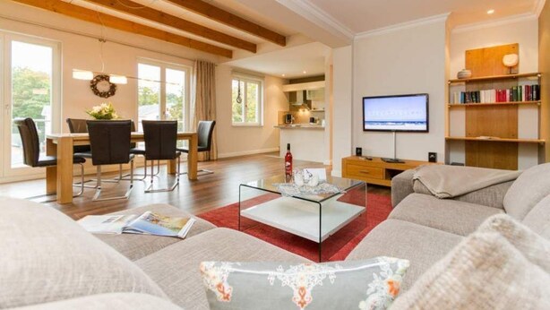 A spacious living room with a large sofa, a glass coffee table, and a television. There is a dining area with chairs and a table, and a kitchen in the background.
