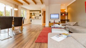 A modern living room with a sofa, a coffee table, and a TV. There is a dining area with chairs and a table. The room has wooden floors and a red rug.