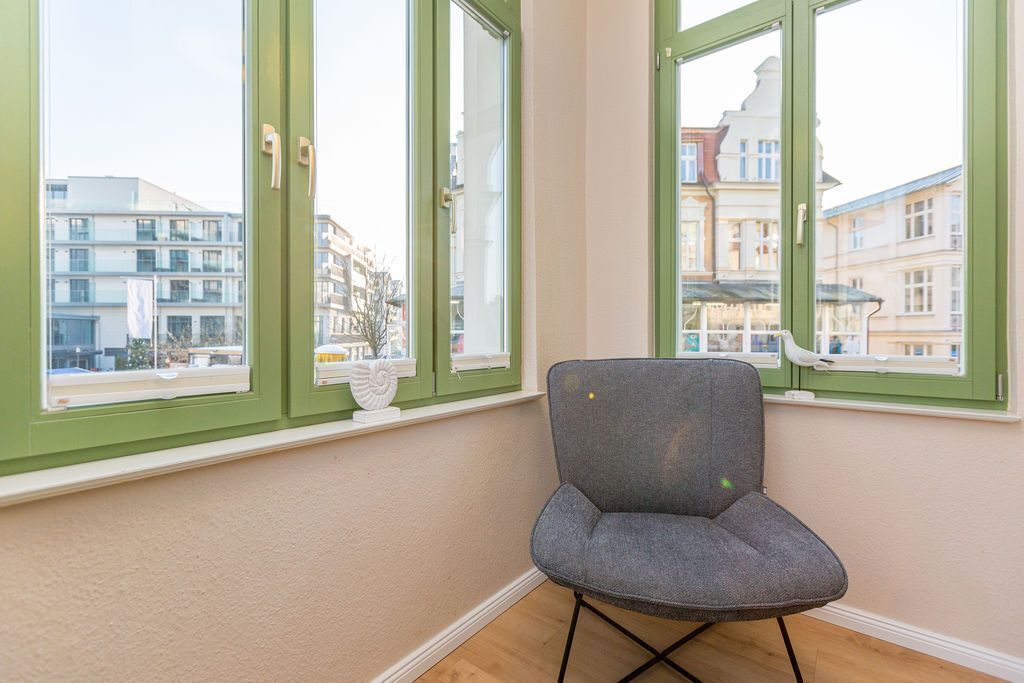 A room with a chair near a window with green frames, a white wall, and a view of buildings outside.