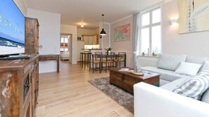 A modern living room with a sofa, coffee table, and TV. There is a dining area with a table and chairs. The room has wooden floors and a large window. - Villa Frisia, app. 30th (Heringsdorf)