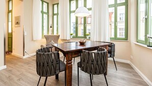 A dining room with a wooden table and six chairs, large windows with green frames, and a hanging light fixture.