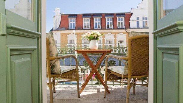 A balcony with wicker chairs and a small table, overlooking a row of buildings.