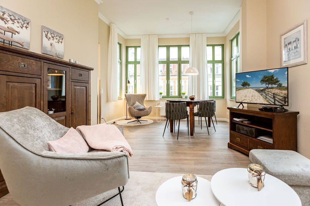 Ein geräumiges Wohnzimmer mit grauem Sofa, Couchtisch und Fernseher auf einem Holzständer. Zwei Sessel und ein Esstisch mit einem Kronleuchter sind ebenfalls vorhanden.