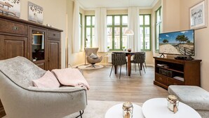 A spacious living room with a grey sofa, a coffee table, and a TV on a wooden stand. There are two chairs and a dining table with a chandelier.