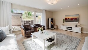 Ein Wohnzimmer mit Sofa, zwei Sesseln, Couchtisch, Kamin, Fernseher und einem großen Fenster.