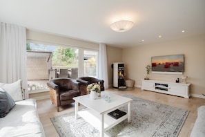 Een woonkamer met een bank, twee fauteuils, een salontafel, een open haard, een televisie en een groot raam.