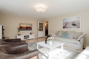 A living room with a white coffee table, brown leather sofa, a beige sofa, a TV on the wall, and a painting of a pier.