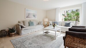 Ein Wohnzimmer mit einer Couch, einem Couchtisch, einer Lampe und einem Fenster mit Blick auf Bäume.