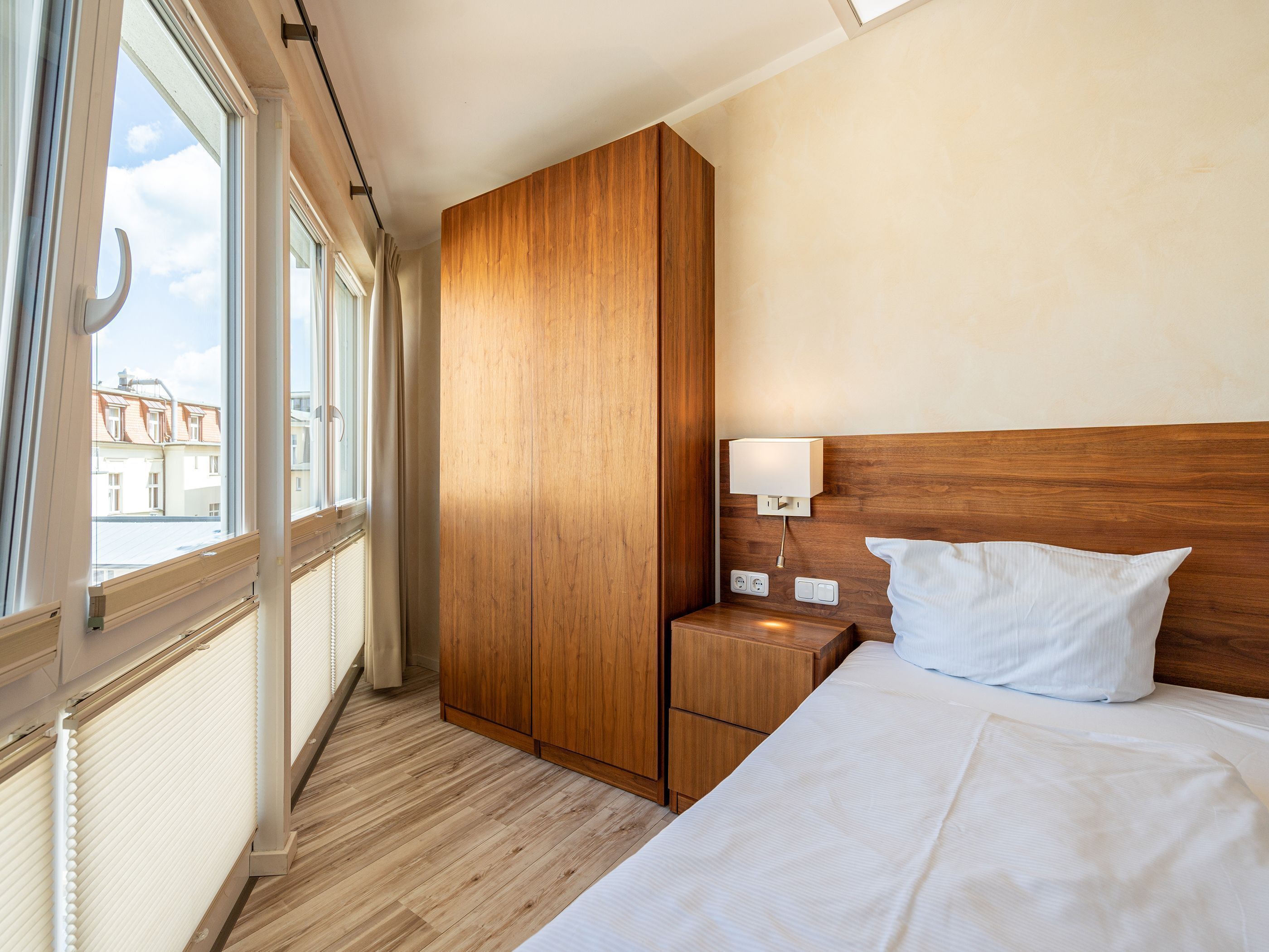 Une chambre d’hôtel avec une armoire en bois, une table de chevet avec une lampe, un lit avec du linge de maison blanc et une fenêtre donnant sur des immeubles.