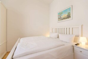 Una cama bien tendida con sábanas blancas, cabecera blanca y mesita de noche con lámpara y control remoto.