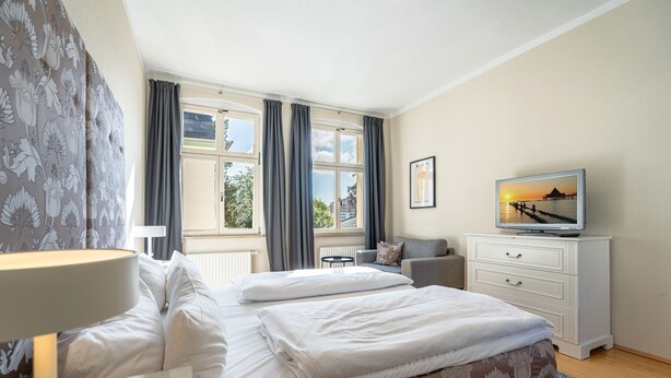 A hotel room with a bed, a TV, a desk, and a window with curtains. - Villa Schlossbauer, App. 06 Remise (Heringsdorf)