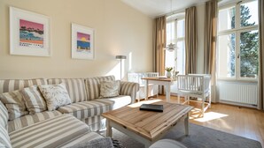 A living room with a striped sofa, a wooden coffee table, a dining table with chairs, and large windows with curtains. - Villa Schlossbauer, App. 06 Remise (Heringsdorf)