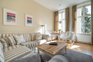 A living room with a striped sofa, a wooden coffee table, a dining table with chairs, and large windows with curtains. - Villa Schlossbauer, App. 06 Remise (Heringsdorf)