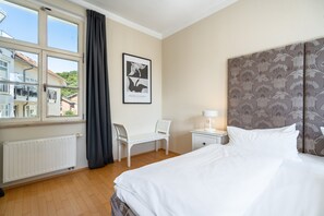 A hotel room with a bed, a desk, a chair, a nightstand, a framed picture on the wall, and a window with curtains. - Villa Schlossbauer, App. 06 Remise (Heringsdorf)