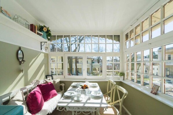 Une salle à manger baignée de lumière naturelle, avec une table dressée pour quatre personnes, de grandes fenêtres et une vue sur l’extérieur.