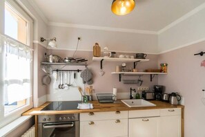 Une cuisine équipée d’une cuisinière, d’un four et de divers ustensiles accrochés au mur. Des étagères au-dessus du plan de travail servent à ranger la vaisselle, et un porte-ustensiles suspendu permet d’y accrocher des casseroles et des poêles.