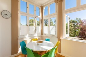 Un comedor con una mesa redonda y sillas coloridas, ventanas amplias con contraventanas blancas, y un reloj en la pared.