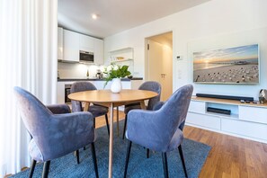 A modern kitchen with white cabinets, a dining area with a round table and four chairs, and a large painting of a beach scene on the wall. - Beach house, App. 08, Germany (Heringsdorf)